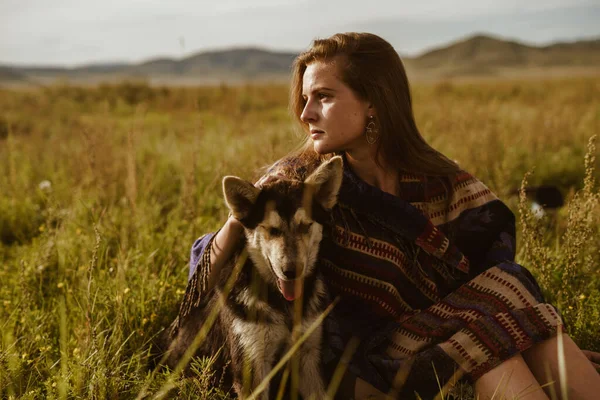 Una chica en una capa de etno se sienta en la hierba de la estepa abrazando la mano de los perros — Foto de Stock
