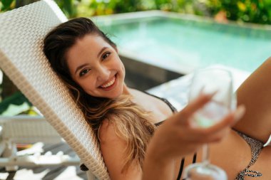 Yanaklarında gamzeleri olan güzel gülümseyen bir kız havuzun kenarında uzanan bir bardağı gerer.