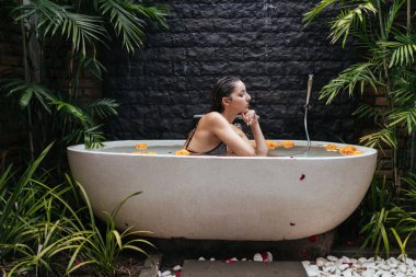 Palmiye ağaçlarıyla çevrili bir kaplıca. Taş duvarın orada su ve çiçek tomurcuklarıyla dolu bir küvet duruyor. Mayo giymiş bir kız çenesine yapışmış duruyor.