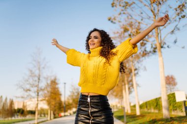 Parlak bir süveter ve deri bir etek giymiş kollarını esneterek sonbahar parkında birine gülümsüyor.