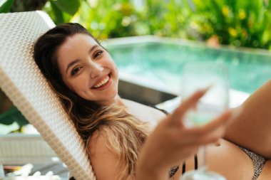 Havuzun kenarında mayo giymiş gülümseyen bir kız yatıyor ve bardağı ileri çekiyor.