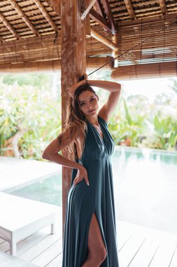 Havuzun kenarındaki ahşap bir terasta, sütundaki bir ağaç gölgesinin altında uzun elbiseli bir kız duruyor ve diğer eliyle saçına dokunuyor.