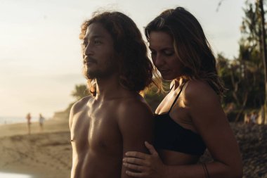 Deniz kenarındaki kumların üzerinde palmiye ağaçlarının arka planında çıplak gövdeli bir adam mesafeye bakar ve arkasından bir kadın tarafından nazikçe sarılır ve ellerini sıvazlar.