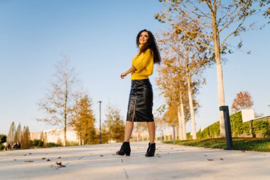 Sonbahar parkındaki pistte parlak sarı kazaklı ve uzun kıvırcık saçlı siyah deri etekli bir kız duruyor.