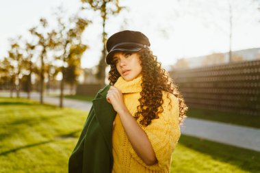 Yeşil gözlü güzel esmer, düşünceli bir şekilde parkın yeşil çimlerinde paltonun omzunun üzerinden süzülüyor.