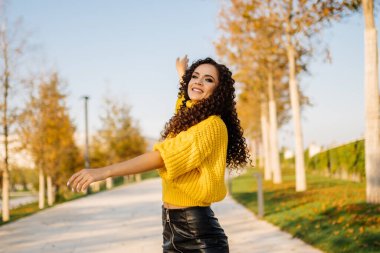 Parlak kazaklı, uzun siyah kıvırcık saçlı bir kız sonbahar ağaçlarıyla çevrili bir parkta dans ediyor..