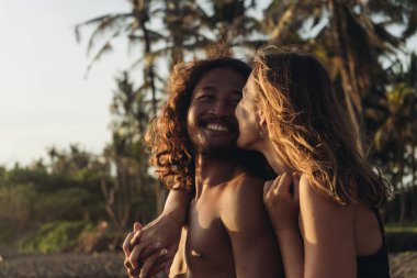 Palmiye ağaçlarının ve kumların arkasında, gülümseyen erkek ve kadınlar birbirlerine bakıp sarılıyorlar.