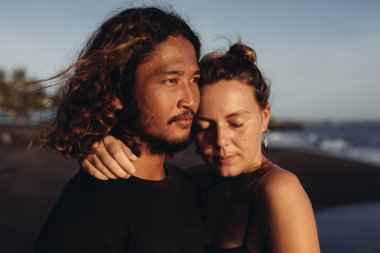 Doğu Asyalı bir adam, kız elini boynundan tutup ona sarılırken ve yanağını bükerek gözlerini kapatırken uzaklığa bakar.