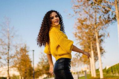 Siyah deri etekli, sarı kazaklı, kolları sonbahar parkında poz veren esmer kadın.