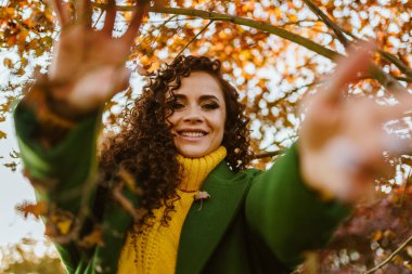 Kız ellerini kameraya çekiyor beyaz dişli büyüleyici gülüşü ile sarı ağaç yapraklarının arka planına doğru