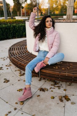 Parktaki bankta tek bacağının altına sıkıştırılmış ve elini uzatmış saçının bir kıvrımını dikkatlice uzaktaki kıza bakıyor.