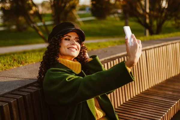 Sitting on a bench in the park and flaunting looks at himself in the phone screen and makes selfies a beautiful girl — Stock Photo, Image