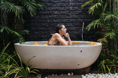 Açık havada, su ve çiçek tomurcuklarıyla çevrili bir kızın oturduğu taş bir duvar ve palmiye yaprağının arka planında bir banyo duruyor.