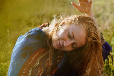 Bir çayırda etno pelerinli bir kız oturuyor başını eğerek saçlarını savuruyor ve elleriyle onları ele geçiriyor.