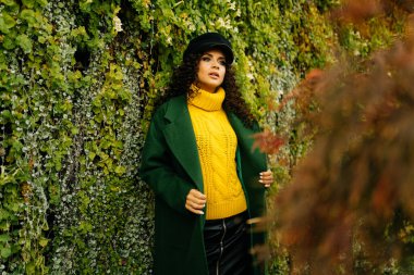 Duvar, arka planda sarmaşıkla çevrili. Şık kıyafetler içinde duran, uzun saçlı, sıra dışı güzellikte bir kız.