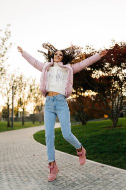 Kalın elbiseli bir kız park yolunda zıplıyor, saçları yanlara saçılmış ve kollarına gülümsüyor.