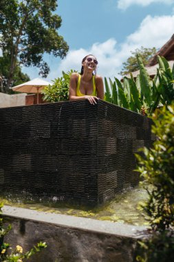 Havuzdan onun tarafına bakan güneş gözlüklü güzel bir esmer, yeşilliğin arka planında.