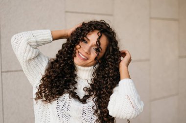 Kıvırcık esmer, neşeli bir gülümsemesi var. Başının kenarına doğru eğilmiş saçlarının arkasını düzleştiriyor.