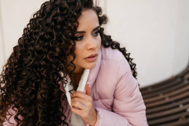 Yakın plan. Kıvırcık esmer, hüzünlü bir bakışla dalgın görünüyor. Hafifçe dolgun dudaklarını açıyor ve el yakalı bir kazak tutuyor.