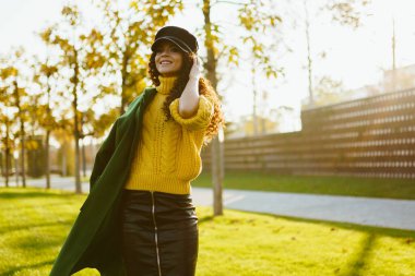 Parktaki yeşil çayırda sarı parlak kazaklı, kıvırcık bir esmer duruyor omzuna yeşil bir ceket atıyor.