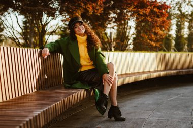 Parkta bankta sonbahar ceketiyle oturuyor ve güzel kıvırcık bir esmer ve büyüleyici bir gülüşü var.