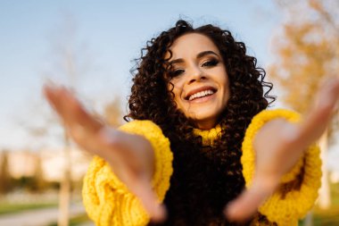 Parlak sarı bir kazak içinde neşeli bir gülümsemesi olan kıvırcık esmer ellerini kameraya çeker.