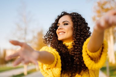 Kıvırcık siyah saçlı, gülümseyen bir kız iki elini de ileri uzatıyor.