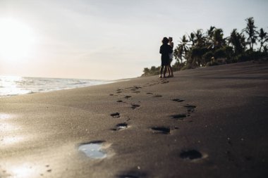 Okyanus kıyısındaki ıslak kumun üzerinde bir iz zinciri var. Uzağa doğru gidiyor. Palmiye ağaçlarının arka planında bir erkek ve bir kadın var.
