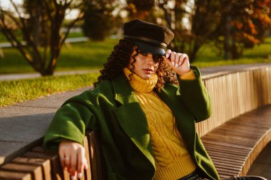 Parktaki uzun ahşap bir bankta sonbahar kıyafetleri giymiş bir kız oturuyor. Elleriyle şapkanın güneşliğine tutunuyor.