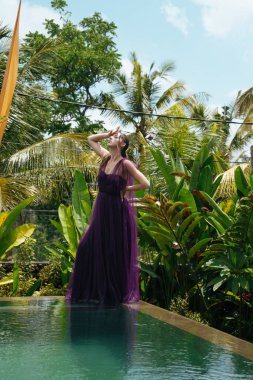 Havuzun kenarında uzun elbiseli bir kız duruyor resim gibi elini başının arkasında tutuyor ve başka bir küçük düşürücü.