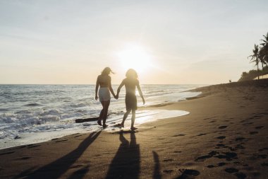 Gün batımının arka planında okyanusun üzerinde su kenarında el ele tutuşan iki erkek ve kadın gider.