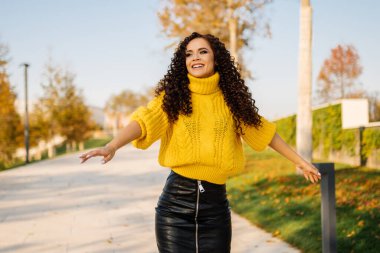 Parlak süveter ve deri etekli bir kız kollarını açmış gülerek sonbahar parkında