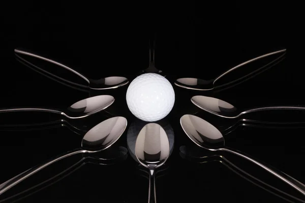 Teaspoons and white golf ball on the black glass table — Stock Photo, Image