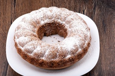 Pudra şekeri serpiştirilmiş halka şeklinde bir kek. Ahşap arka planda beyaz bir tabakta ev yapımı kek..