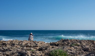 Genç bir kız, güneşli ve rüzgarlı bir günde sahilde telefonuyla dalgaların fotoğraflarını çekiyor..
