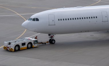 Önlük taşıyıcısı, uluslararası bir havaalanının hava sahasında modern, dar gövdeli beyaz bir yolcu uçağını çekiyor..