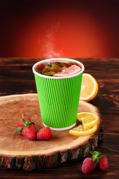 Tea Mockup. Fruit tea with raspberries and lemon in green paper cup on wooden background. Space for text