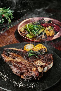 Izgara sebzelerle servis edilen T-Bone bifteği siyah yuvarlak bir tabakta yeşil, baharat ve sebzelerden oluşan bir arka planda, bakır bir çarşaf üzerinde. Kapat.