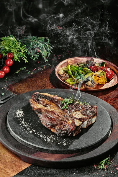 T-Bone steak podávaný s grilovanou zeleninou na černém kulatém talíři na pozadí zeleně, koření a zeleniny, na měděném plechu. zavřít — Stock fotografie