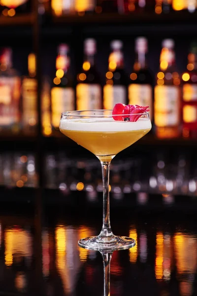 Elegant female yellow cocktail with white foam, decorated with pink flower petal on dark table