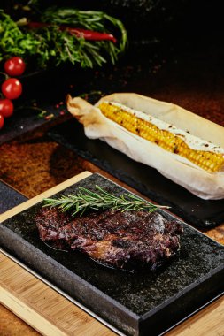 Mayalanmış biftek. Siyah bir tabakta biberiyeli biftek ve yeşil, baharatlı ve taze sebzeli ızgara mısır. Bakır bir çarşaf üzerinde. Kapat. seri fotoğraf