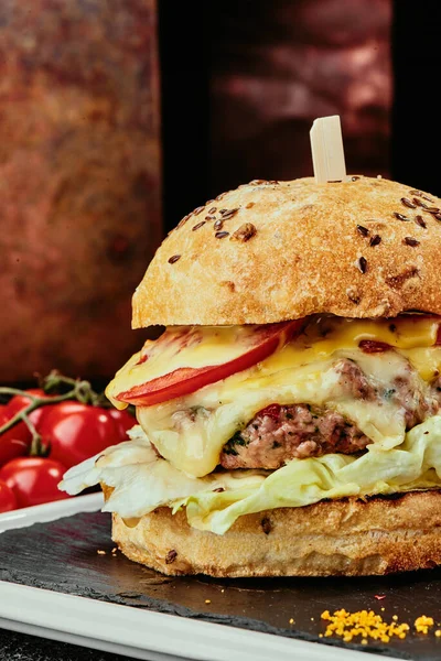 Nagy hamburger lédús szelettel, sajttal, paradicsommal, salátával egy fekete tányéron, rézlemezek hátterén. Zárj be! makroszintű — Stock Fotó