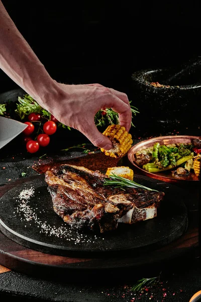 T-Bone steak podávaný s grilovanou zeleninou na černém kulatém talíři na pozadí zeleně, koření a zeleniny, na měděném plechu. zavřít — Stock fotografie