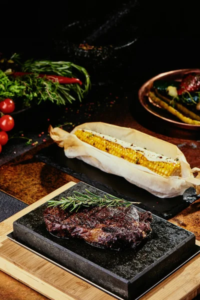 Kvašený steak. Steak s rozmarýnem na černém talíři podávaný s grilovanou kukuřicí na pozadí zeleně, koření a čerstvé zeleniny, na měděném plechu. Zblízka. série fotografií — Stock fotografie