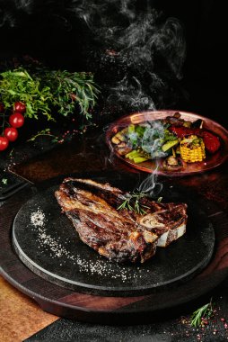 Izgara sebzelerle servis edilen T-Bone bifteği siyah yuvarlak bir tabakta yeşil, baharat ve sebzelerden oluşan bir arka planda, bakır bir çarşaf üzerinde. Kapat.