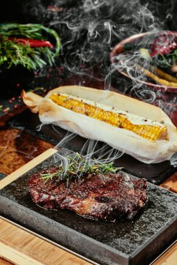 Mayalanmış biftek. Siyah bir tabakta biberiyeli biftek ve yeşil, baharatlı ve taze sebzeli ızgara mısır. Bakır bir çarşaf üzerinde. Kapat. seri fotoğraf