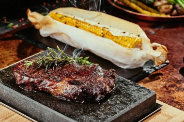 Mayalanmış biftek. Siyah bir tabakta biberiyeli biftek ve yeşil, baharatlı ve taze sebzeli ızgara mısır. Bakır bir çarşaf üzerinde. Kapat. seri fotoğraf