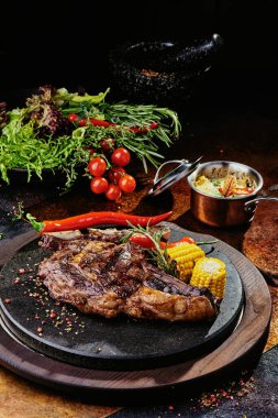 Izgara sebzelerle servis edilen T-Bone bifteği siyah yuvarlak bir tabakta yeşil, baharat ve sebzelerden oluşan bir arka planda, bakır bir çarşaf üzerinde. Kapat.