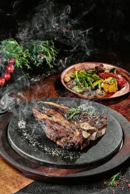 Izgara sebzelerle servis edilen T-Bone bifteği siyah yuvarlak bir tabakta yeşil, baharat ve sebzelerden oluşan bir arka planda, bakır bir çarşaf üzerinde. Kapat.