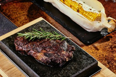 Mayalanmış biftek. Siyah bir tabakta biberiyeli biftek ve yeşil, baharatlı ve taze sebzeli ızgara mısır. Bakır bir çarşaf üzerinde. Kapat. seri fotoğraf